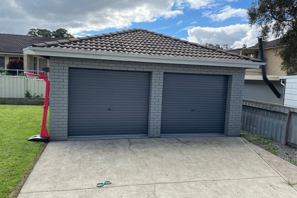 A detached grey brick double garage with two dark grey roller doors and a tiled roof.