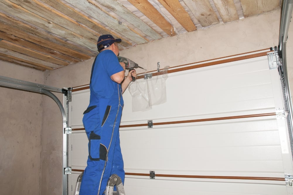 A man in blue using a power drill to install a white sectional garage door in a concrete room with a wooden ceiling.