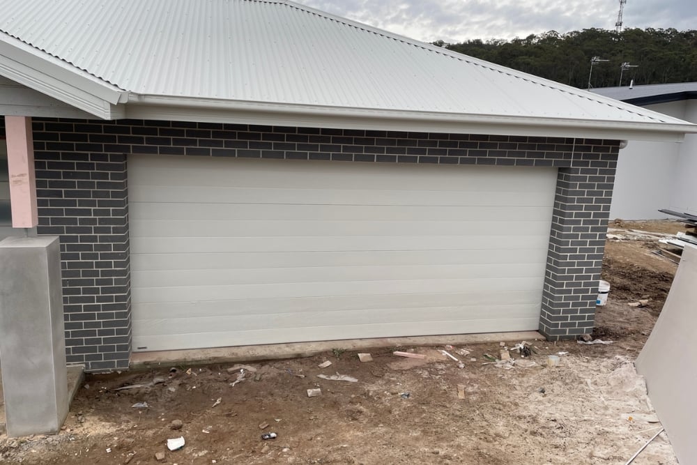 A wide, light-coloured sectional garage door set in a dark grey brick wall under a corrugated metal roof.