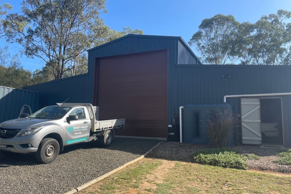 A silver pickup truck for Coast-to-Valley Garage Doors is parked on a gravel driveway in front of a large dark-blue metal industrial shed featuring a maroon roller door.