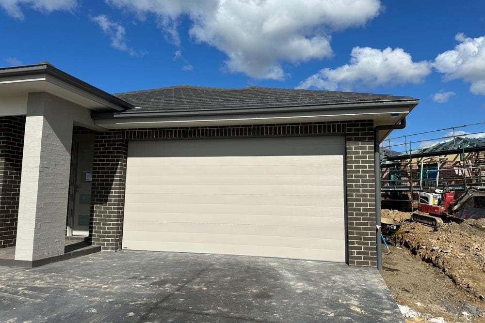 A modern beige sectional garage door installed in a dark brick house under a clear blue sky.