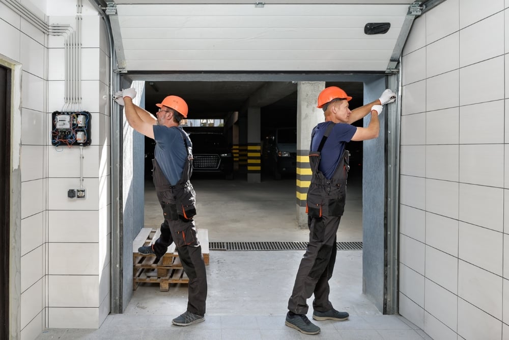 Two technicians in orange hard hats and work overalls performing maintenance on the tracks of an open sectional garage door