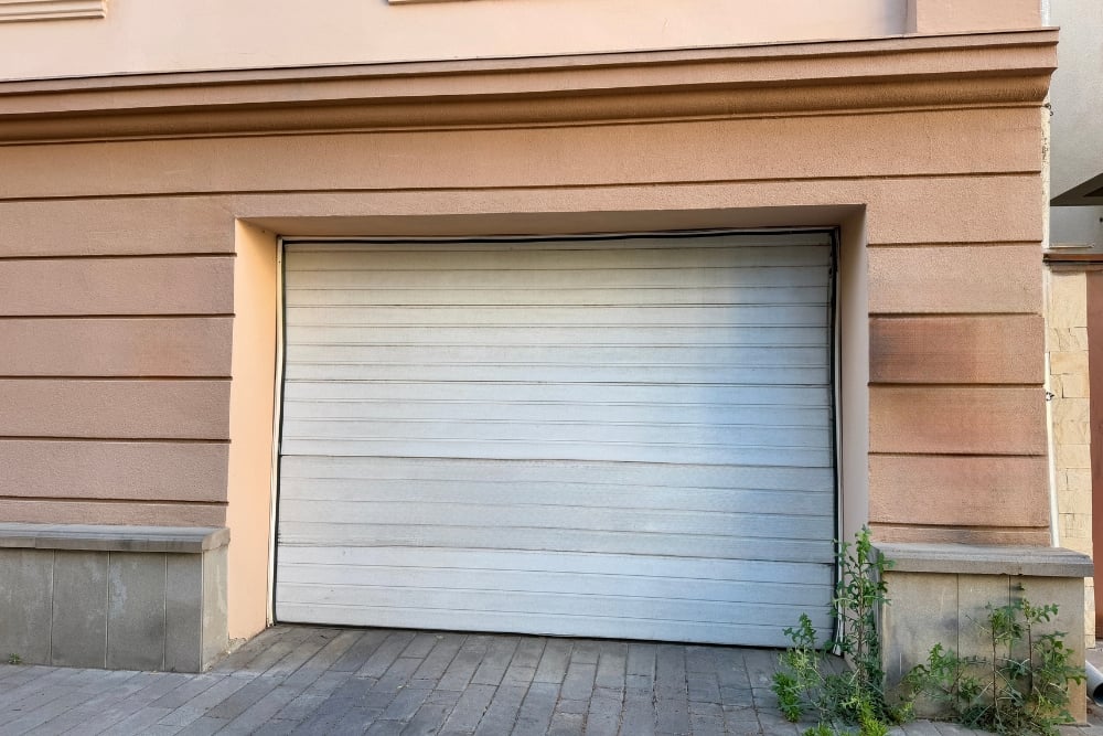 A closed white sectional garage door that is visibly misaligned and damaged at the bottom, appearing to have been struck by a vehicle.