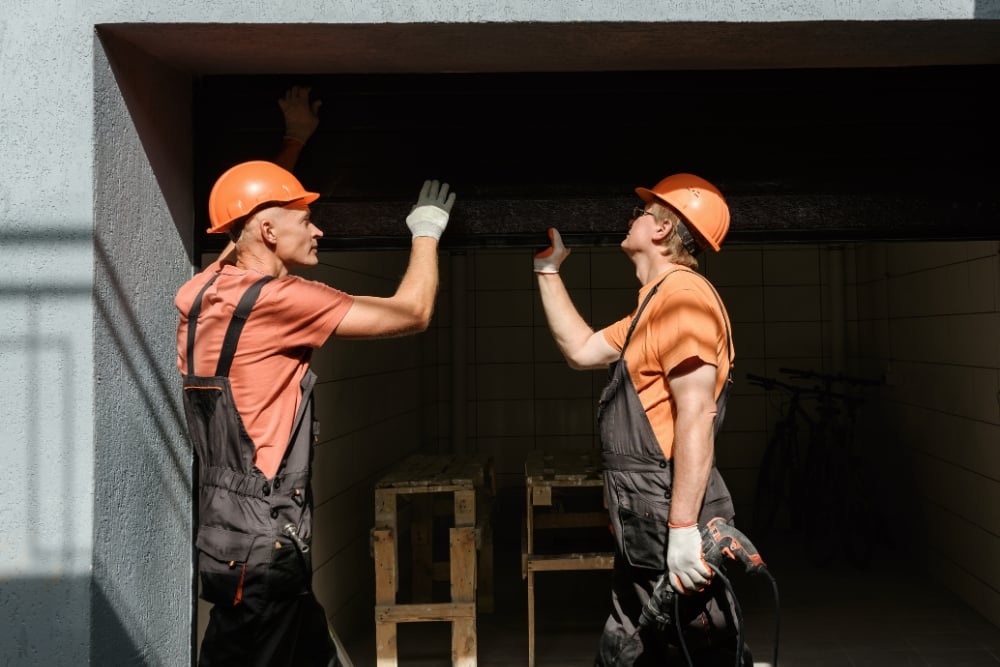 Two technicians wearing orange hard hats and work overalls installing a dark roller garage door at a multi-unit complex.