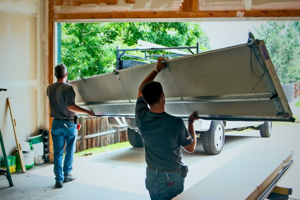 Two technicians manually lifting a large, single garage door panel into place during a replacement at a residential property.