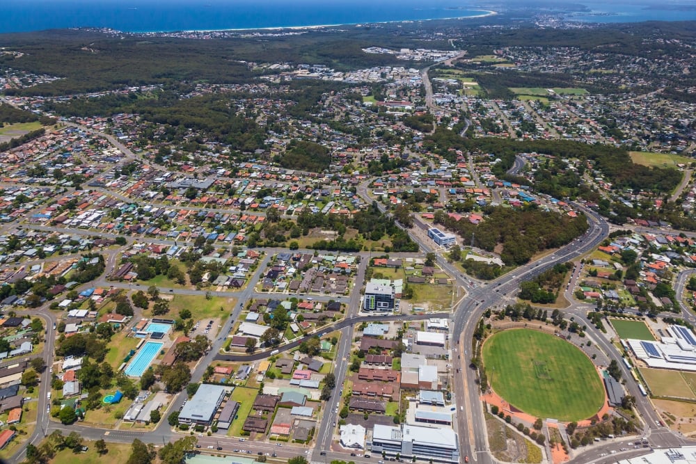 Aerial view of a suburban neighborhood showing houses, a large green sports oval, main roads, and the coastline in the far background.