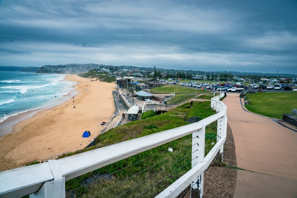A sandy beach coastline meeting turquoise ocean waves under an overcast sky, looking past a white-railed coastal walkway toward a seaside parking lot and a hilly town backdrop.