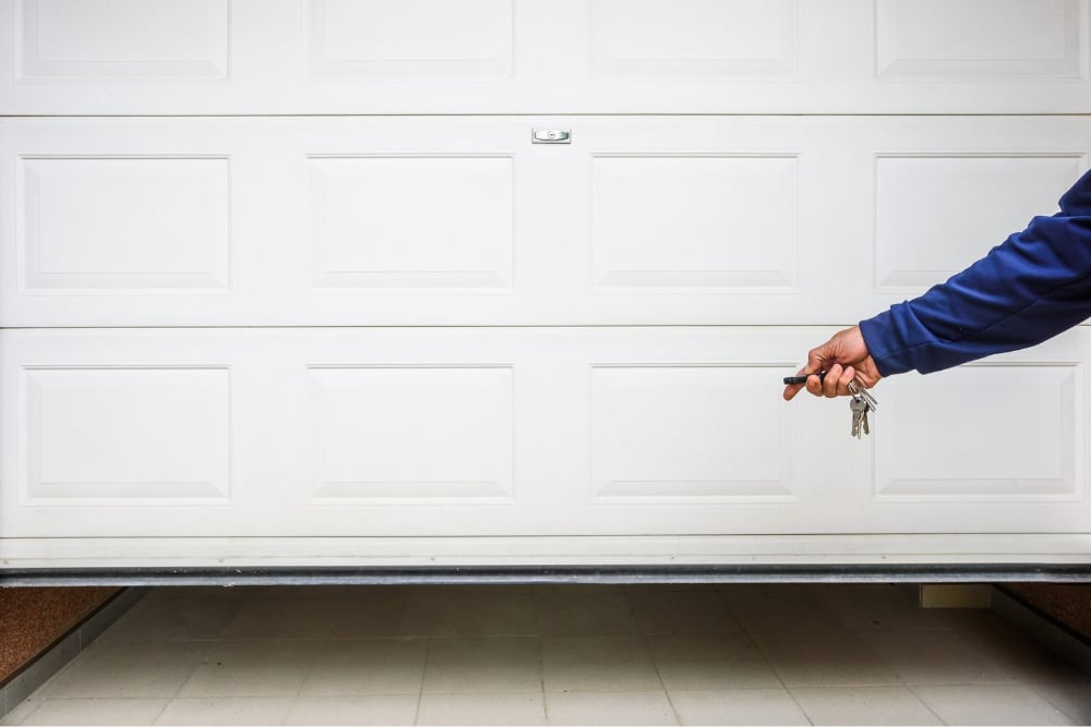 A close-up of a person's hand holding a remote and keys to open a white, paneled sectional garage door that is slightly open at the bottom.