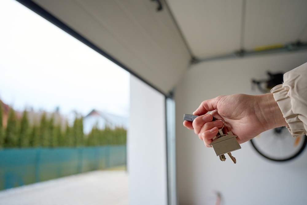 A close-up of a hand holding a keychain remote inside a garage, pressing a button to operate an open sectional garage door.