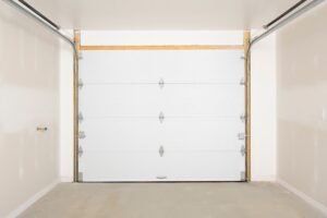 Interior view of an empty garage showing a closed, white sectional garage door with metal hinges and horizontal tracks under unfinished beige walls and a concrete floor.