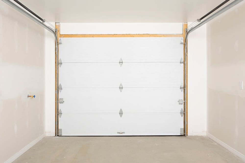 Interior view of an empty garage showing a closed, white sectional garage door with metal hinges and horizontal tracks under unfinished beige walls and a concrete floor.
