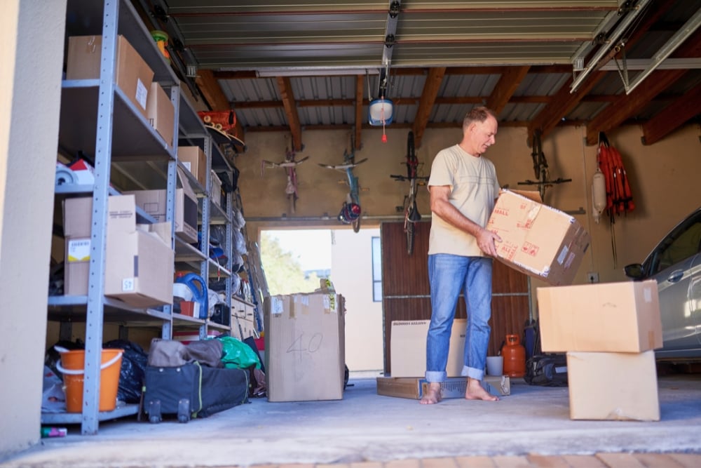 A man moving cardboard boxes to organise or clear a cluttered garage filled with storage shelving and bicycles hanging on the back wall.