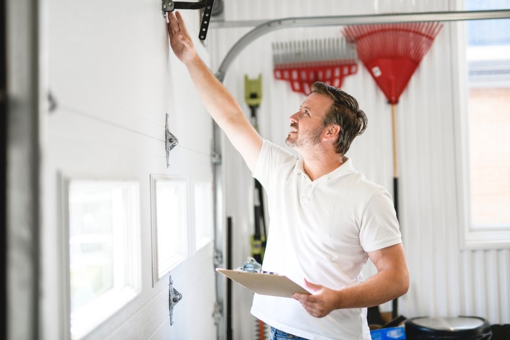 A technician holding a clipboard inspects the hinges and hardware on the inside of a white sectional garage door.