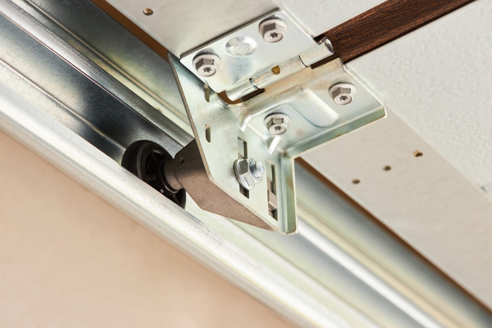 A close-up of a garage door roller mechanism sitting inside a metal track, attached to a door panel hinge with bolts.