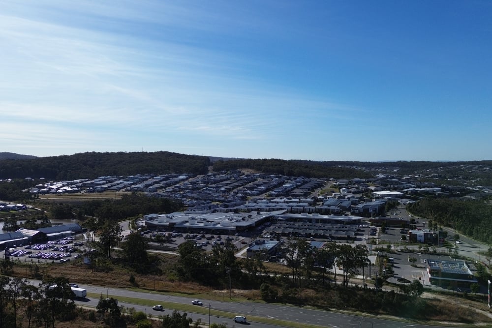 A sprawling suburban residential development and a commercial shopping center with parking lots, set against a backdrop of forested hills under a clear blue sky.