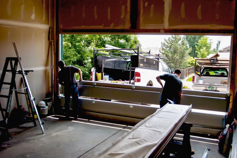 An unfinished garage showing two workers installing a white sectional garage door.